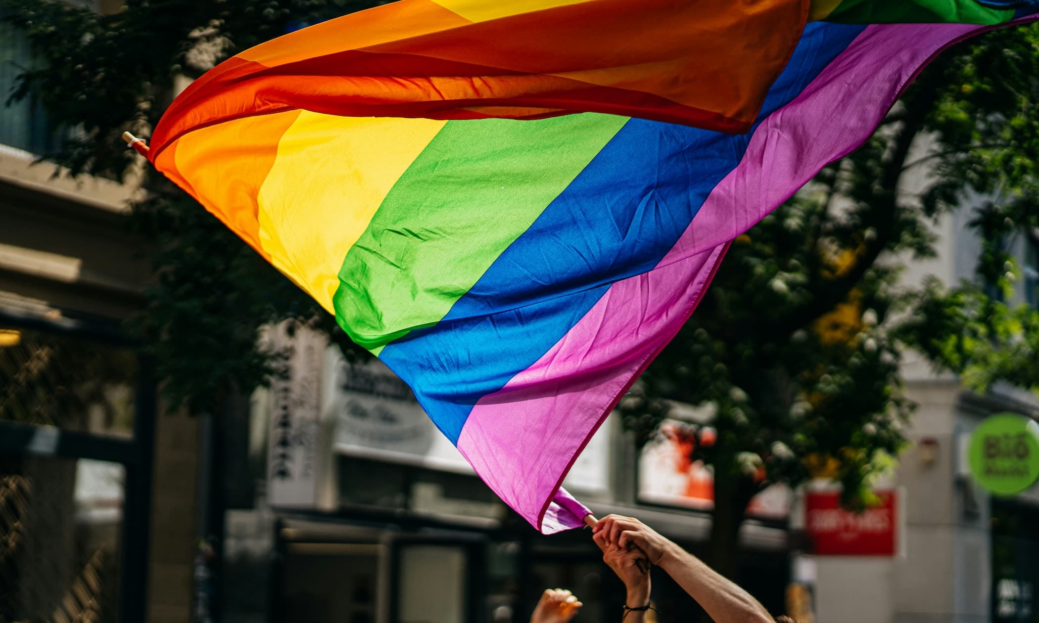 Regenbogenfahne, die von Händen im Wind geschwenkt wird. Hintergrund: eine Straße mit Gebäuden und Bäumen.
