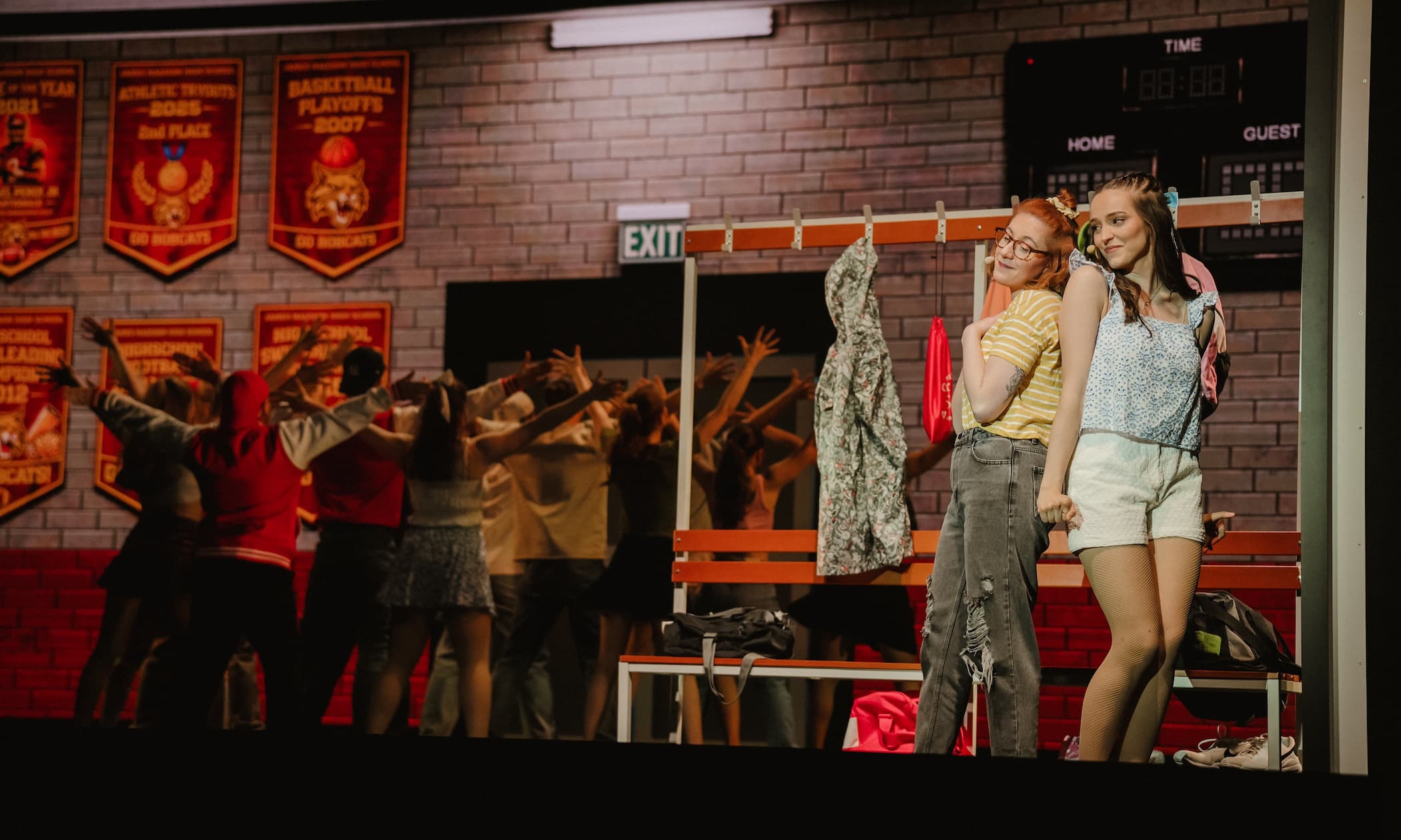 Szenenfoto aus dem Musical The Prom: Zwei Darstellerinnen stehen im Vordergrund an einer Umkleidebank und lächeln sich an. Im Hintergrund tanzt ein Ensemble mit erhobenen Armen vor einer Kulisse, die eine Highschool-Sporthalle mit Backsteinwand und Sport-Bannern darstellt.