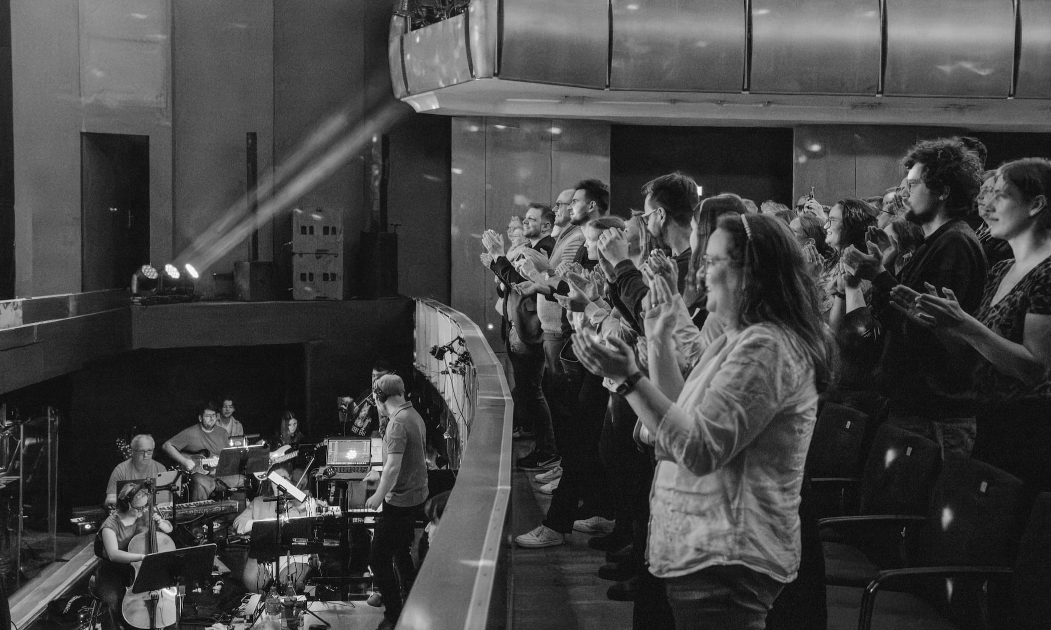 Publikum auf Balkon applaudiert stehend. Links unten spielt Live-Orchester im Graben. Schwarz-Weiß-Foto im Theatersaal.