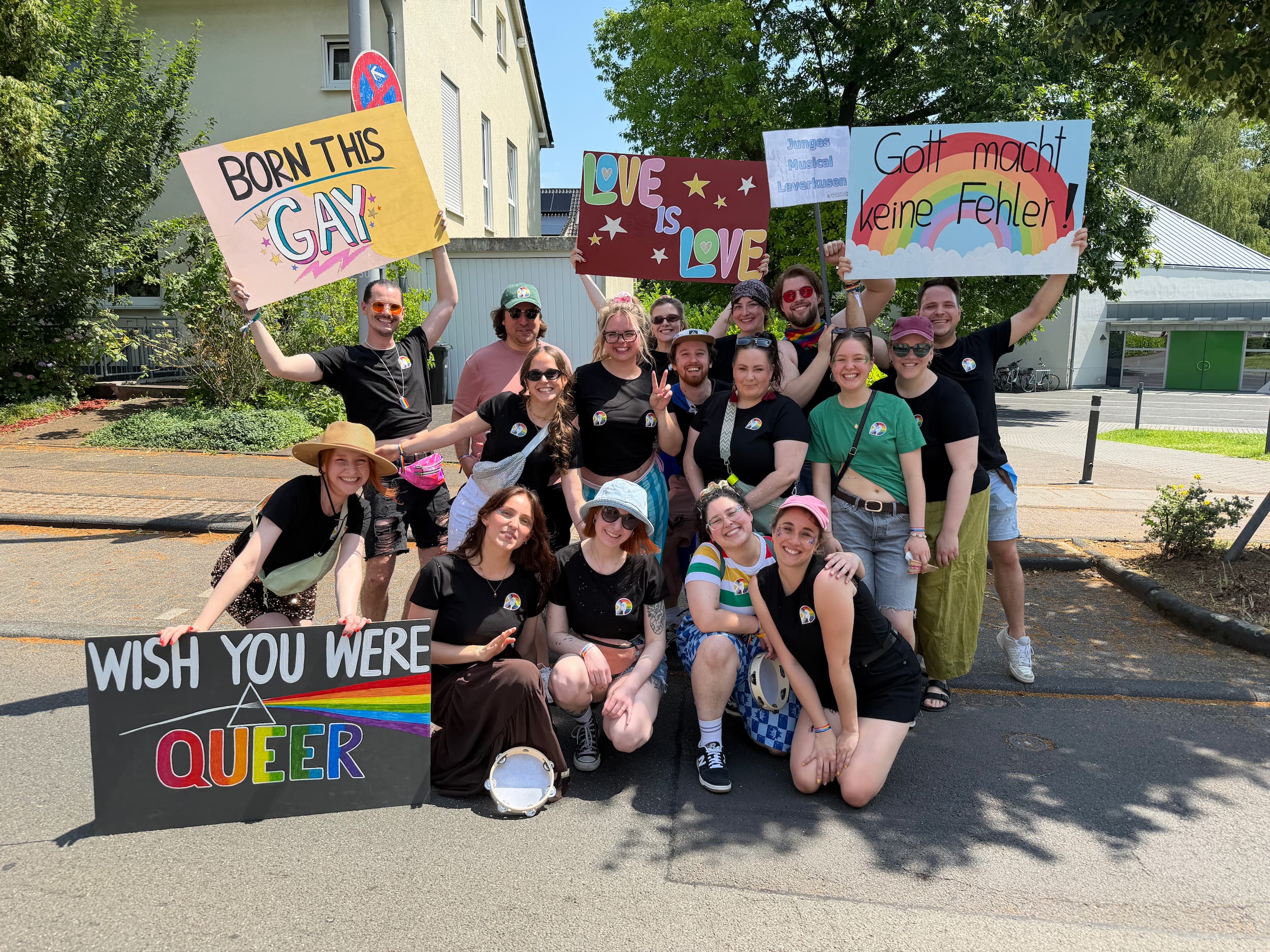 Eine Gruppe von über einem Dutzend lächelnden Menschen posiert im Freien auf einer Straße bei einer Pride-Demonstration und hält selbstgestaltete Schilder mit bunten Regenbogen-Motiven und Slogans wie „Born This Gay“, „Love is Love“, „Gott macht keine Fehler!“ und „Wish You Were Queer“ hoch.