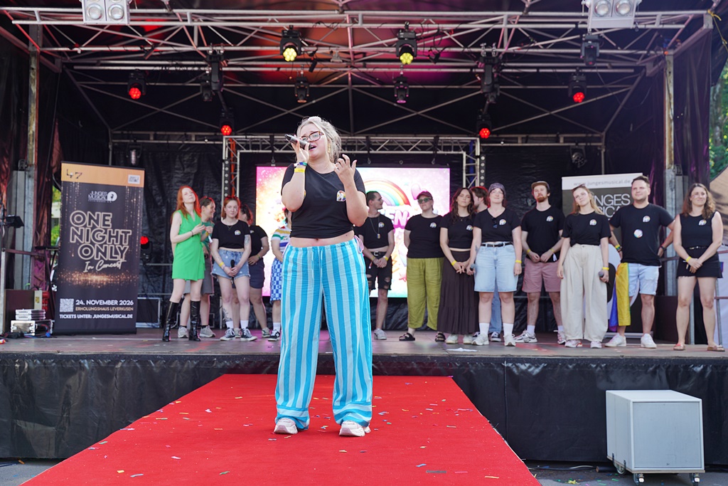 Eine Sängerin performt mit einem Mikrofon auf einem roten Teppich vor einer Festivalbühne, während im Hintergrund eine Gruppe junger Menschen im Halbkreis steht und zusieht.